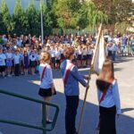 Uczniowie w mundurkach stoją na zewnątrz w rzędach, a trzech z nich trzyma flagę podczas apelu szkolnego.