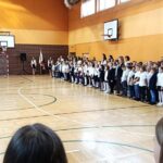 Apel szkolny w sali gimnastycznej; uczniowie i pracownicy szkoły w strojach uroczystych stoją w rzędach, twarzą do siebie.
