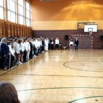 Uczniowie i nauczyciele stoją w rzędach w sali gimnastycznej podczas uroczystości; w tle widać kosz do koszykówki.