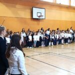 Uczniowie i nauczyciele stoją w sali gimnastycznej, ustawieni w szeregach na apelu szkolnym lub uroczystości.