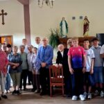Grupa osób w różnym wieku stoi razem w pomieszczeniu z elementami wystroju o charakterze religijnym i roślinami.