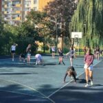 Nastolatki ćwiczą na zewnętrznym boisku do koszykówki, otoczonym drzewami i budynkami mieszkalnymi.