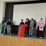 Uczniowie stojący na scenie i trzymający kolorowe tabliczki w kształcie strzałek.