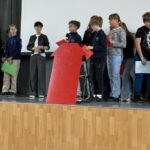 Uczeń przemawia do mikrofonu, stojąc za dużą czerwoną papierową strzałką na scenie wraz z innymi osobami.