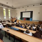 Studenci siedzący na trybunach uniwersyteckiej auli, zwróceni twarzą w stronę sceny z ekranem projekcyjnym.