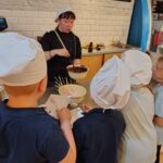 Kobieta pokazuje dzieciom, jak maczać smakołyki w czekoladzie; wszyscy mają na głowach czapki kucharskie i znajdują się w kuchni.