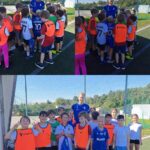 Trener piłki nożnej stoi wraz z grupą dzieci na zewnętrznym boisku sportowym.