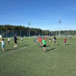 Trener i grupa dzieci grających w piłkę nożną na trawiastym boisku.