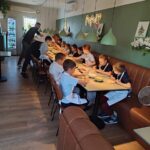 Grupa dzieci w fartuszkach przygotowująca pizzę przy stoliku w restauracji wraz ze swoim instruktorem.