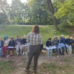 Nauczyciel stoi przed grupą uczniów siedzących na ławce w parku z notesami z klipsem w rękach.