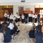 Grupa uczniów stoi na scenie i występuje przed siedzącą publicznością w dużej sali.