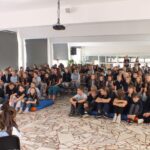 Duża grupa uczniów siedzących na podłodze w szkolnej auli i słuchających występu gitarowego.