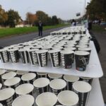 Rzędy markowych papierowych kubków wypełnionych wodą, stojących na stole podczas imprezy plenerowej.