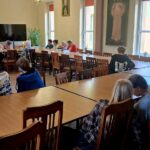 Ludzie siedzący przy długich drewnianych stołach w sali, na ścianach której wiszą obrazy o tematyce religijnej.