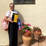Kobieta trzymająca podręcznik do języka hiszpańskiego przed szyldem Malaca Instituto, przy którym stoją doniczki z kwiatami.