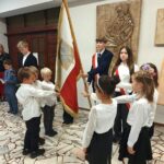 Grupa uczniów w oficjalnych mundurkach szkolnych biorąca udział w uroczystym salutowaniu flagi.