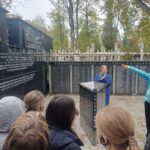 Przewodnik przemawia do grupy uczniów przed pomnikową ścianą z czarnego granitu, na której wyryto nazwiska.