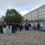 Grupa studentów stoi na brukowanym dziedzińcu i ogląda tablice informacyjne poświęcone historii.