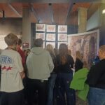 Grupa osób ogląda zdjęcia oraz duży historyczny plakat wystawiony w galerii.