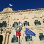 Flagi Malty i Unii Europejskiej powiewające przed bogato zdobionym kamiennym budynkiem.
