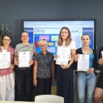 Siedem osób stoi przed ekranem, a każda z nich trzyma w ręku certyfikat Eti.