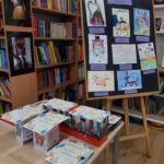Wystawa prac plastycznych uczniów na sztalugach oraz kilka dyplomów na stole w bibliotece.