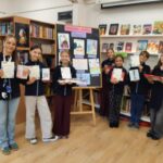 Siedmioro uczniów stojących w bibliotece z dyplomami i nagrodami w rękach przed tablicą ogłoszeń.